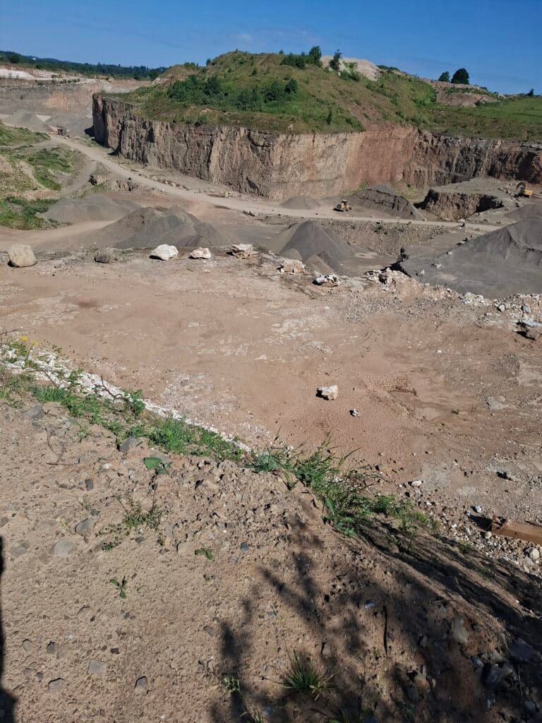 Open-pit quarry with piled gravel, rocky cliff walls, and distant trucks under a clear blue sky.