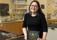 Smiling woman scientist in a black shirt at a lab bench, holding a framed chalk drawing of a winged skeleton.