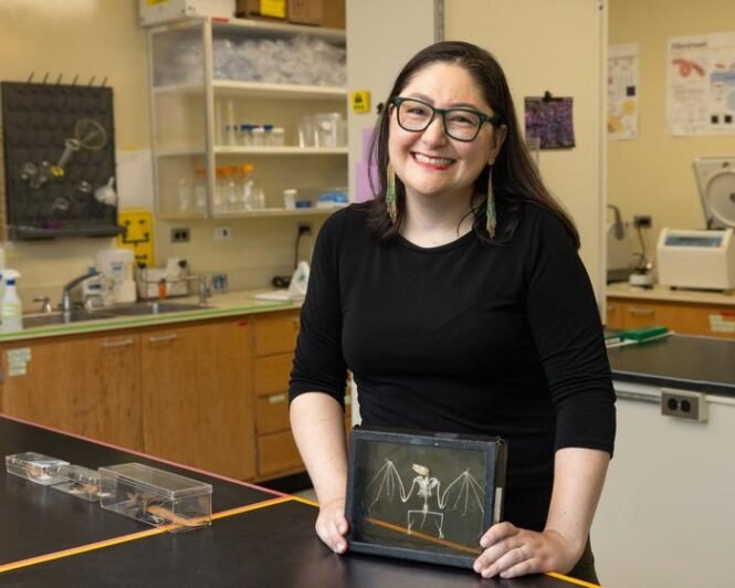 Low-Res_Hannah-1jpg | Pugnalom Smiling woman scientist in a black shirt at a lab bench, holding a framed chalk drawing of a winged skeleton.