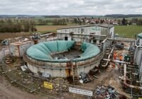 Industrial biogas plant with a large circular digester, rusted metal structures and two workers in orange safety gear nearby.