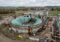 Industrial biogas plant with a large circular digester, rusted metal structures and two workers in orange safety gear nearby.
