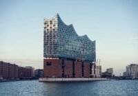 Elbphilharmonie concert hall with a wavy glass facade rising from a brick base by Hamburg harbor, reflected in the water.5c2 The iconic Hamburg building with a glassy, curved roof on the riverfront.5c2