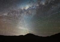 Silhouette of a person on a hill at night, with a bright light casting rays across a star-filled sky and streaking meteor trails.