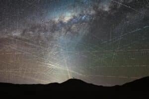 Silhouette of a person on a hill at night, with a bright light casting rays across a star-filled sky and streaking meteor trails.