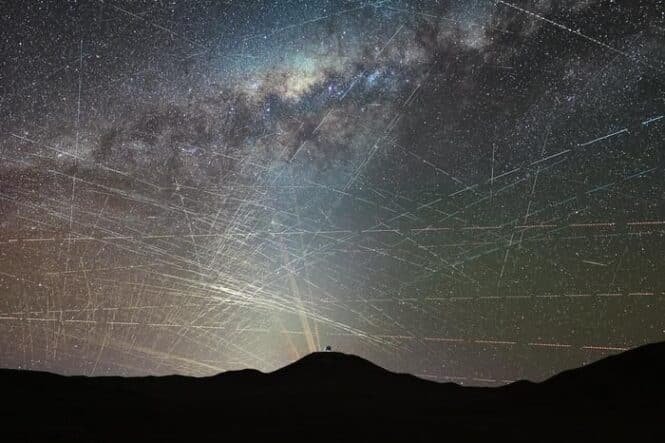 Silhouette of a person on a hill at night, with a bright light casting rays across a star-filled sky and streaking meteor trails.