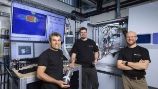 Three engineers in a high-tech lab pose by a workstation with large monitors displaying data and a complex machine behind them.