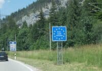 Blue EU border sign for Germany ('Bundesrepublik Deutschland') on a rural road, with a car and forested hillside in the background.