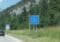 Blue EU border sign for Germany ('Bundesrepublik Deutschland') on a rural road, with a car and forested hillside in the background.