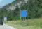 Blue EU border sign for Germany ('Bundesrepublik Deutschland') on a rural road, with a car and forested hillside in the background.