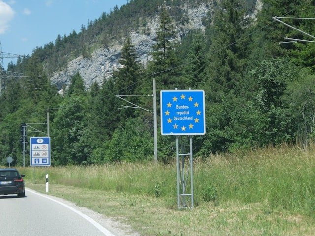 Blue EU border sign for Germany ('Bundesrepublik Deutschland') on a rural road, with a car and forested hillside in the background.