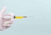 Gloved hand holding a syringe filled with yellow liquid and a long needle against a pale background. The image implies a medical injection is prepared.