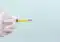 Gloved hand holding a syringe filled with yellow liquid and a long needle against a pale background. The image implies a medical injection is prepared.