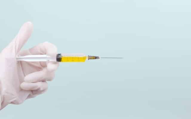 Gloved hand holding a syringe filled with yellow liquid and a long needle against a pale background. The image implies a medical injection is prepared.