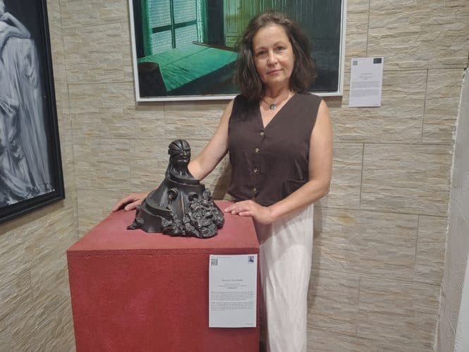 Woman in a brown sleeveless blouse posing with a dark abstract sculpture on a red pedestal in a gallery.
