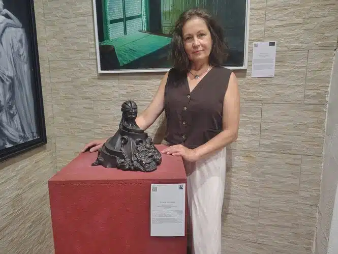 Woman in a brown sleeveless blouse posing with a dark abstract sculpture on a red pedestal in a gallery.