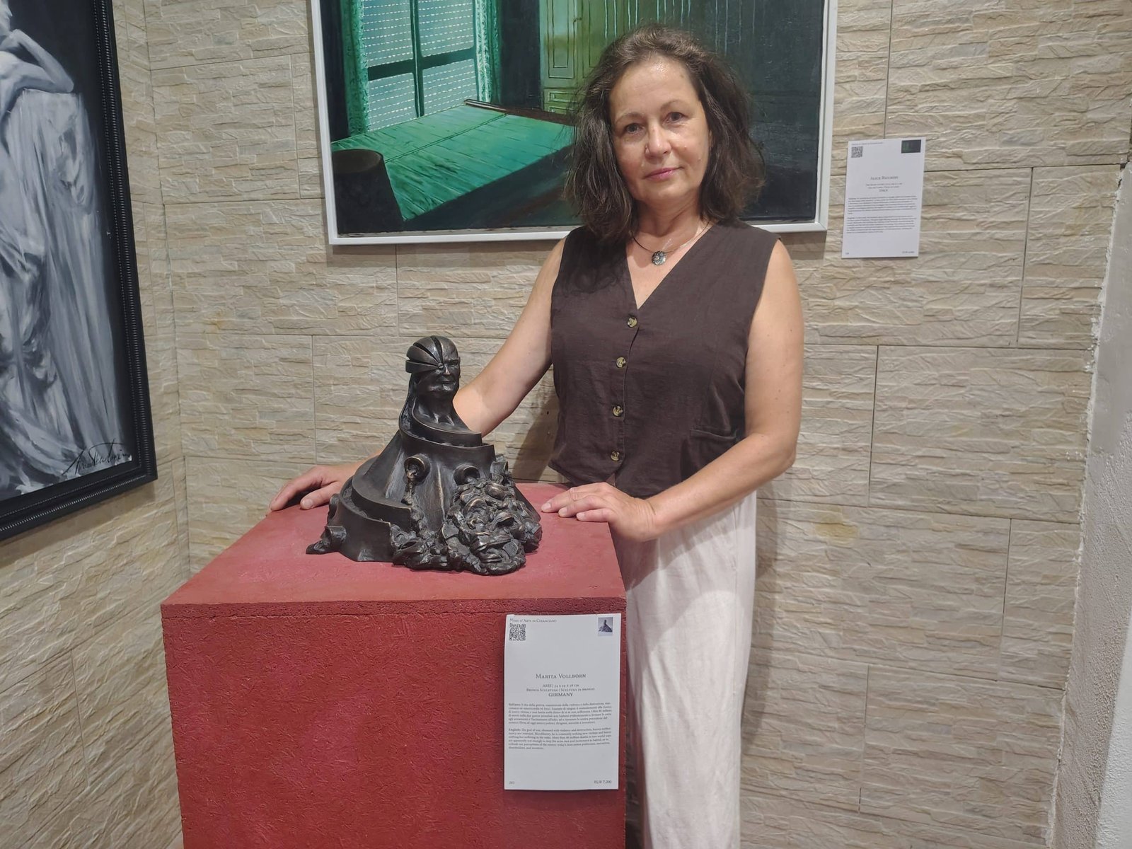 Woman in a brown sleeveless blouse posing with a dark abstract sculpture on a red pedestal in a gallery.