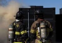 Two firefighters in full turnout gear with SCBA tanks stand side by side, facing a smoky black structure at a training or incident scene.