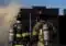 Two firefighters in full turnout gear with SCBA tanks stand side by side, facing a smoky black structure at a training or incident scene.