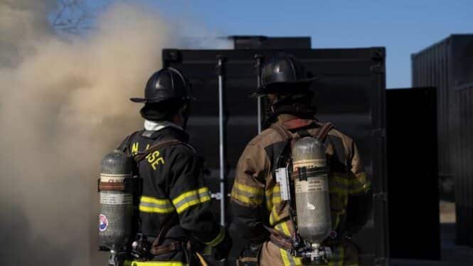 Two firefighters in full turnout gear with SCBA tanks stand side by side, facing a smoky black structure at a training or incident scene.