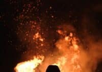 Person silhouetted against a large bonfire at night, sparks flying upward from the flames around them.