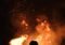 Person silhouetted against a large bonfire at night, sparks flying upward from the flames around them.
