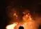 Person silhouetted against a large bonfire at night, sparks flying upward from the flames around them.