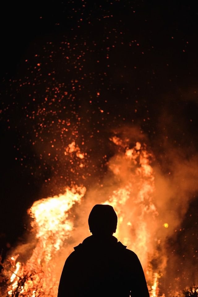 Person silhouetted against a large bonfire at night, sparks flying upward from the flames around them.