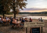 People sit at long wooden tables in an outdoor biergarten by a river at sunset, under trees; a sign reads 'Zum Schwan - Biergarten' and a chalkboard says 'Letzte Badetage'.