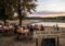 People sit at long wooden tables in an outdoor biergarten by a river at sunset, under trees; a sign reads 'Zum Schwan - Biergarten' and a chalkboard says 'Letzte Badetage'.