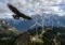 A large eagle soars over a mountain ridge with a row of wind turbines in the distance.