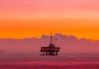 Offshore oil rig standing on stilts in the calm sea at sunset with a vivid orange and pink sky.