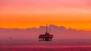 Offshore oil rig standing on stilts in the calm sea at sunset with a vivid orange and pink sky.