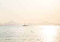 A small sailboat on calm water at sunset, with hazy mountains in the distance and a bright reflection on the sea