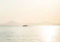 A small sailboat on calm water at sunset, with hazy mountains in the distance and a bright reflection on the sea