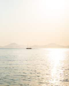 A small sailboat on calm water at sunset, with hazy mountains in the distance and a bright reflection on the sea