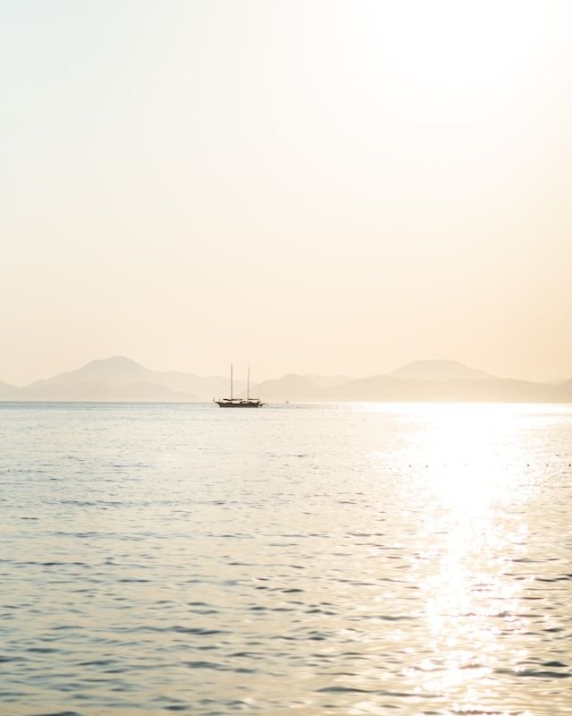 A small sailboat on calm water at sunset, with hazy mountains in the distance and a bright reflection on the sea