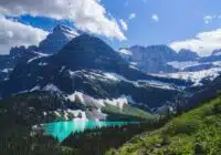 Turquoise alpine lake nestled among evergreen forests and snow-capped mountains under a blue sky.