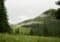 Mountain meadow with evergreen forest, low clouds, and a foggy hillside above a few distant buildings