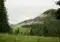 Mountain meadow with evergreen forest, low clouds, and a foggy hillside above a few distant buildings