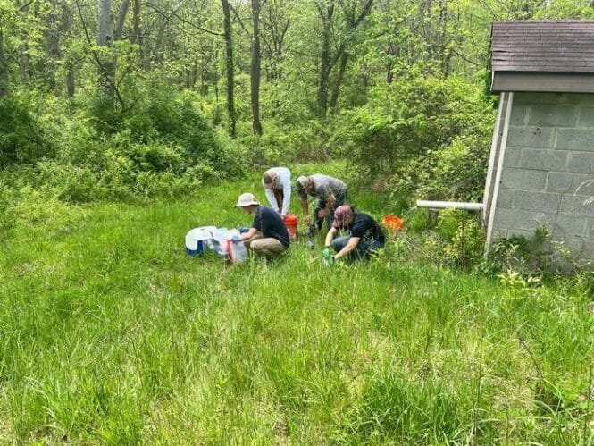 Group of people crouching in tall grass near a small shed, collecting samples or doing fieldwork with coolers and bags nearby.