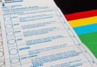 Close-up of a German ballot paper showing party rows (CDU, SPD, FDP, GRÜNE, DIE LINKE) with selection circles, beside a colorful flag.