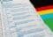 Close-up of a German ballot paper showing party rows (CDU, SPD, FDP, GRÜNE, DIE LINKE) with selection circles, beside a colorful flag.