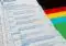 Close-up of a German ballot paper showing party rows (CDU, SPD, FDP, GRÜNE, DIE LINKE) with selection circles, beside a colorful flag.
