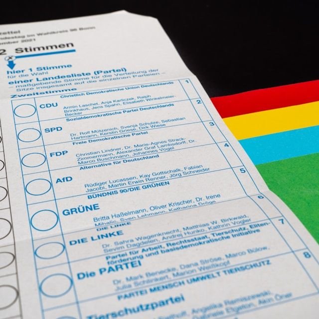 Close-up of a German ballot paper showing party rows (CDU, SPD, FDP, GRÜNE, DIE LINKE) with selection circles, beside a colorful flag.