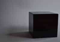 A matte black cube sits on a gray surface with a soft shadow to its left in a neutral studio setting.