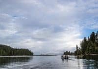 A small boat with people gliding on calm water between forested shores under a cloudy sky.