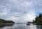 A small boat with people gliding on calm water between forested shores under a cloudy sky.