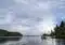 A small boat with people gliding on calm water between forested shores under a cloudy sky.