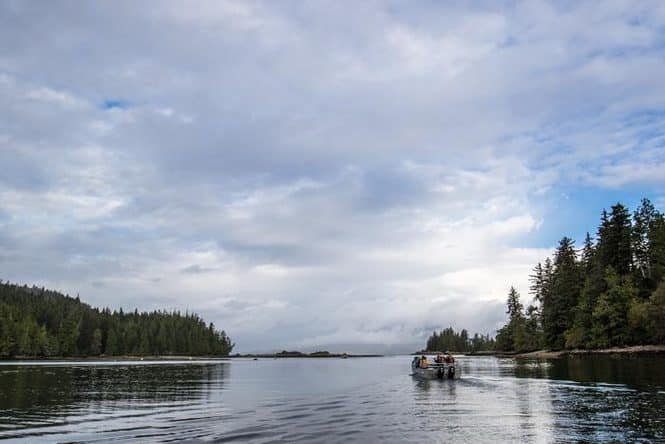 A small boat with people gliding on calm water between forested shores under a cloudy sky.