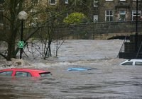 Severe urban flooding: cars partially submerged in brown floodwater on a street, with a bridge and buildings in the background.